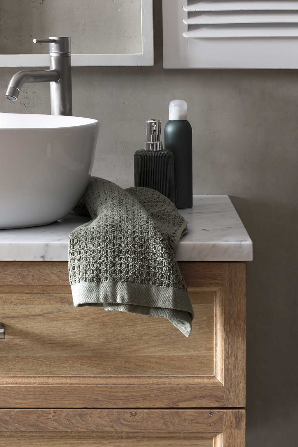 Close-up van een houten badkamermeubel met een marmeren blad, een witte ronde waskom en een moderne roestvrijstalen kraan, versierd met een donkergroene zeepdispenser en een luxe gebreide handdoek