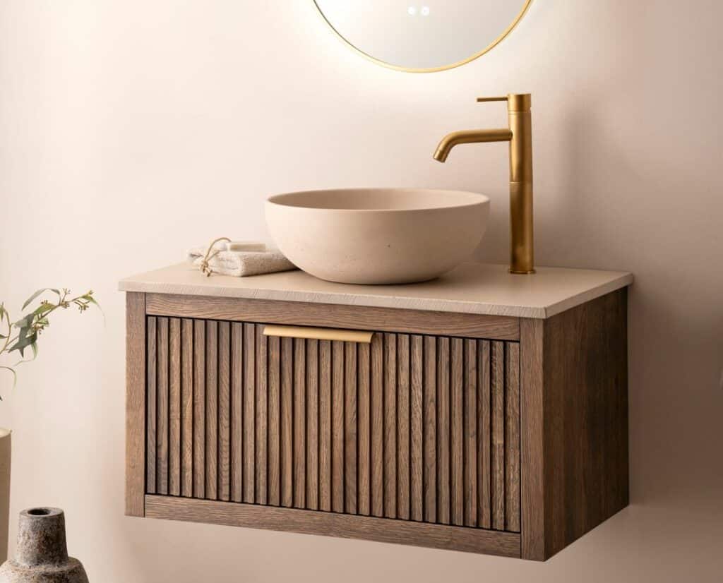 Badkamermeubel van donker hout met gestreept design, voorzien van een beige waskom en een elegante gouden kraan, tegen een zachte beige muur met een ronde spiegel en sfeervolle decoratie.
